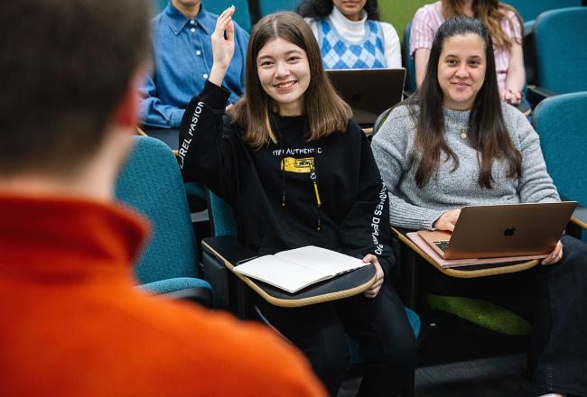 Two Simmons undergraduate students in a class. One of them is raising their hand.