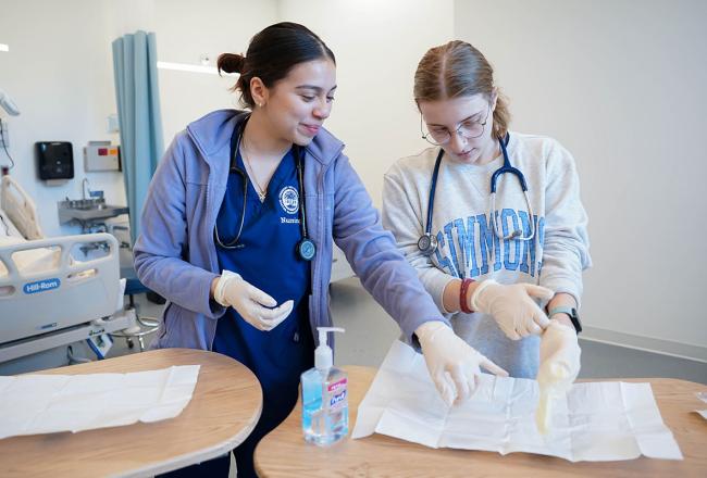 Two Simmons nursing students wearing white gloves and reviewing an instruction guide that is open on a table