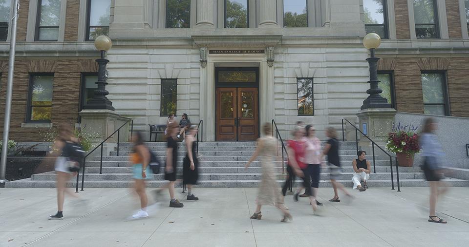 Various people walking by MCB main entrance. One person sitting on steps.