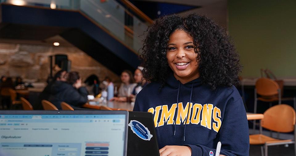 Student in Simmons sweatshirt smiling while on laptop in the Fens