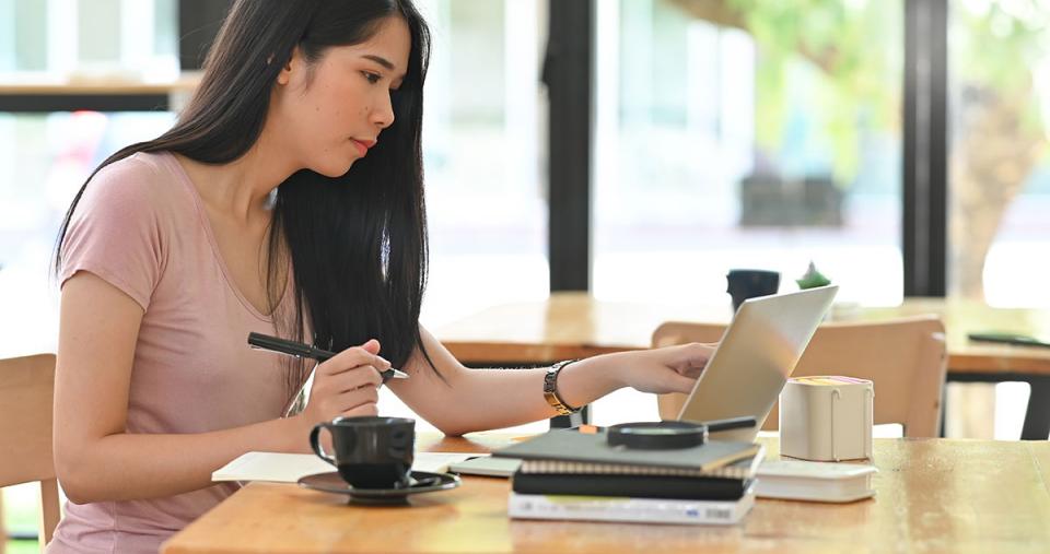 An online student sitting at a table near windows and pointing to a laptop screen on a desk