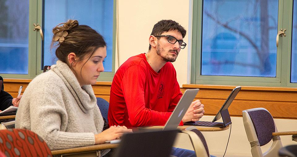 Two students in classroom at desks. One looking down at a laptop - the other has an tablet with a keyboard and is looking forward.