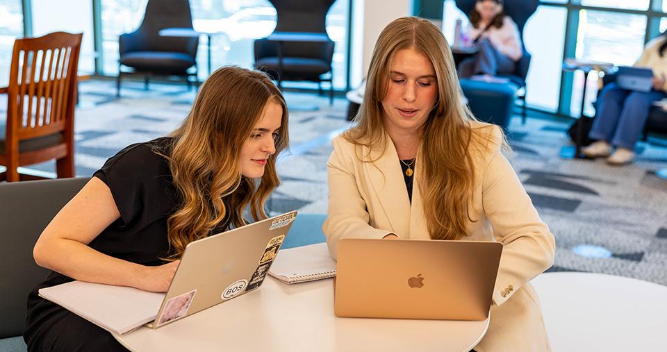Two students working together on laptops in library